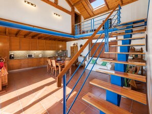 A modern kitchen with wooden cabinets, a dining area with a table and chairs, and a blue staircase leading to a loft bedroom area. - Country House by the Sea (Viana do Castelo)
