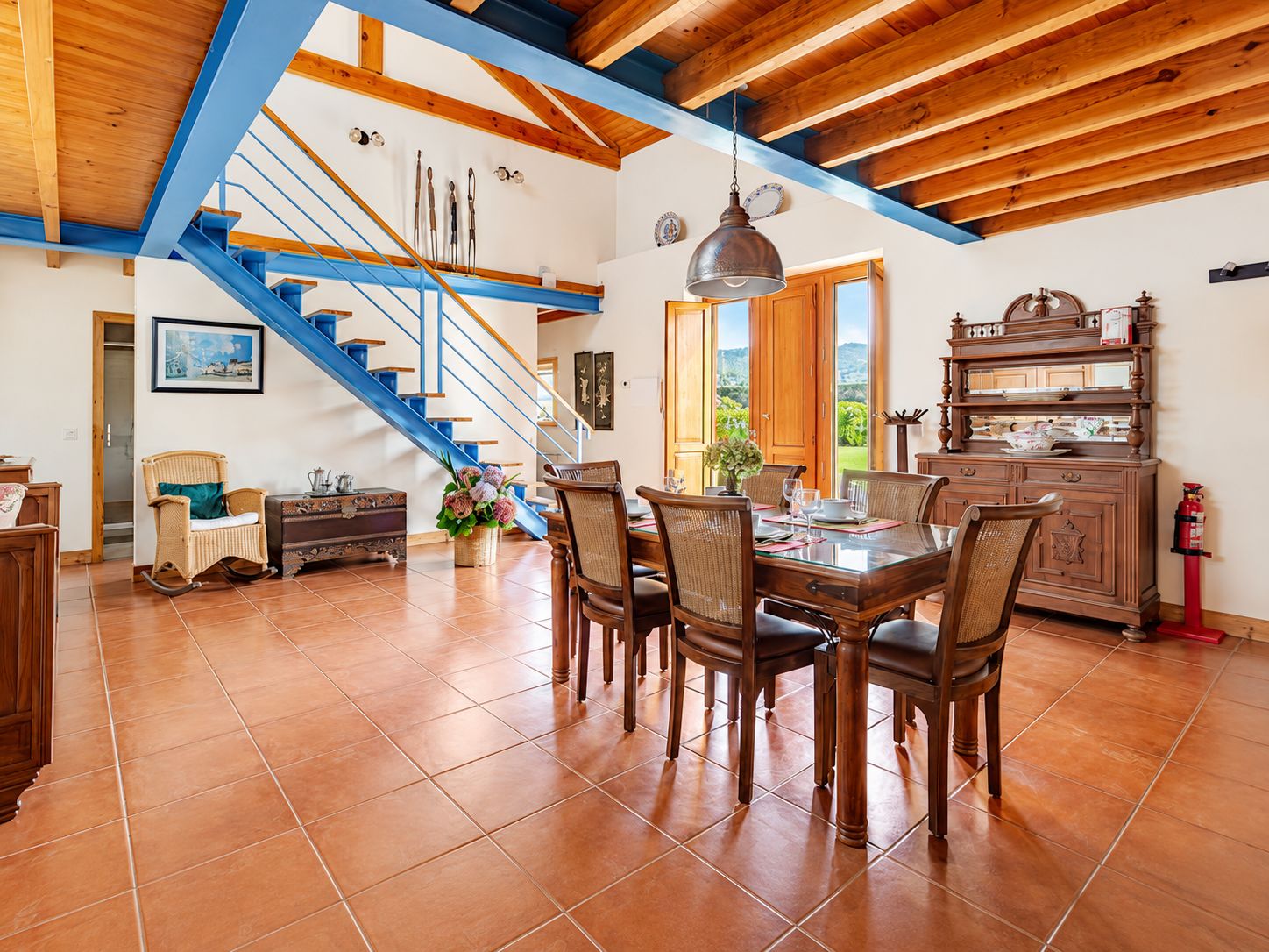 A spacious dining area with a wooden table and chairs, a staircase with blue railings, and a view of the outdoors through a large window.