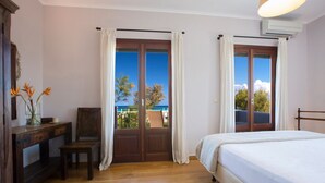 A bedroom with a wooden floor, a bed, a desk with a chair, and a view of the sea through large windows.