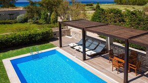 A swimming pool with a wooden deck and lounge chairs, surrounded by greenery and a view of the sea.