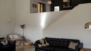 Un salon moderne avec un canapé en cuir noir, une table basse en bois et un lampadaire. Un escalier mène à un niveau supérieur où se trouvent un mur sombre et une fenêtre.