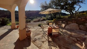 Un espace de restauration en plein air, avec une table et des chaises sous une pergola, entouré d’un paysage naturel et de murs en pierre.