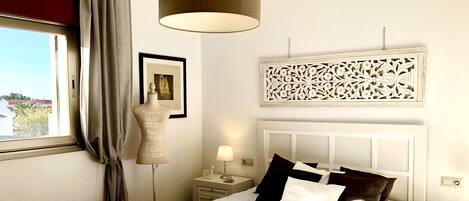 A bedroom with a bed, bedside tables, a lamp, a window with curtains, and a decorative wall piece.