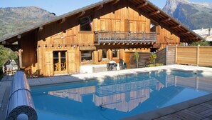 Una baita in legno con un balcone, una piscina e montagne sullo sfondo.