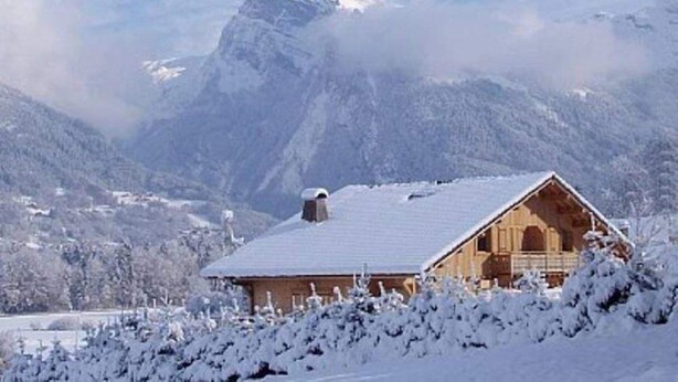Un chalet in legno con tetto spiovente, circondato da alberi innevati e montagne.
