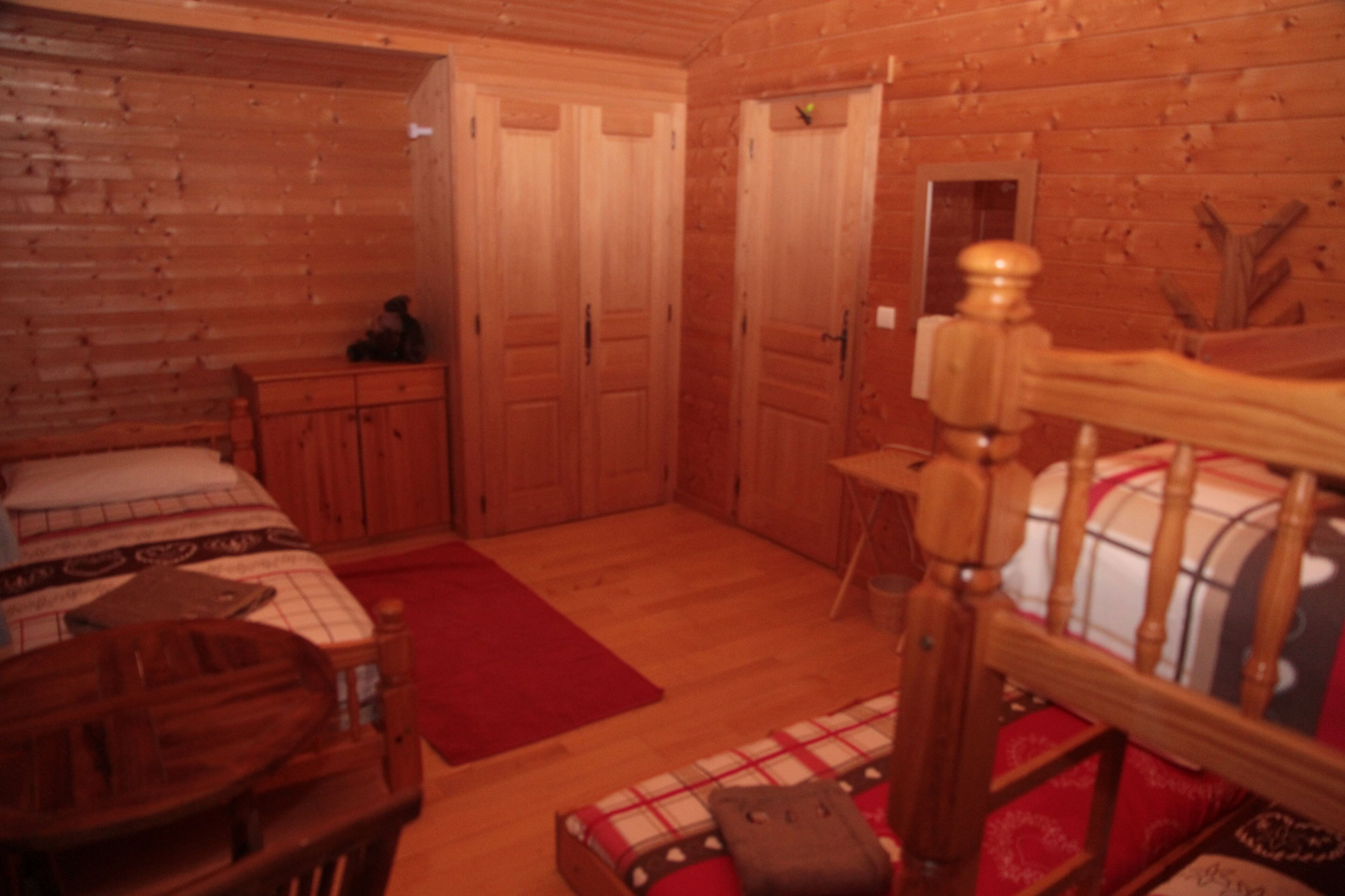 A wooden cabin interior with a bed, a wooden chair, a small table, and a red rug.