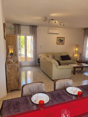 Een woonkamer met een bank, een eettafel gedekt voor twee personen, en uitzicht door een glazen deur op een buitenruimte.