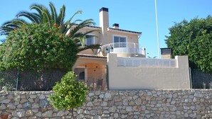 Una villa de estilo mediterráneo con un muro de piedra, una palmera y un cielo azul despejado.