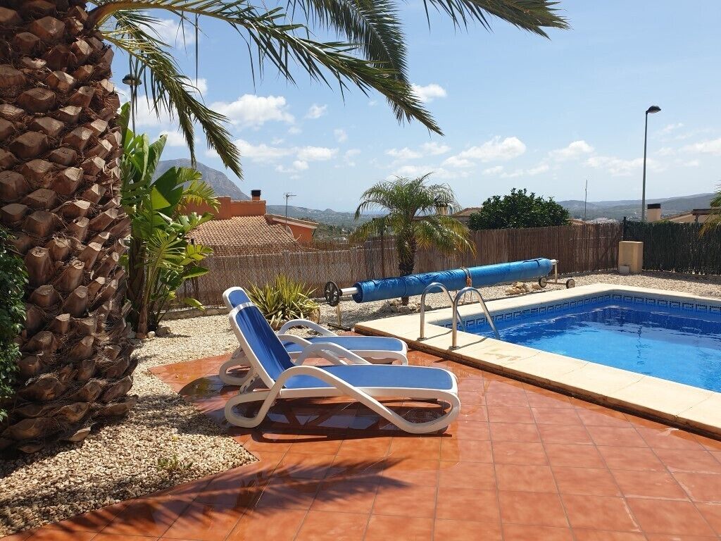 A swimming pool with two lounge chairs, a palm tree, and a mountain view in the background.