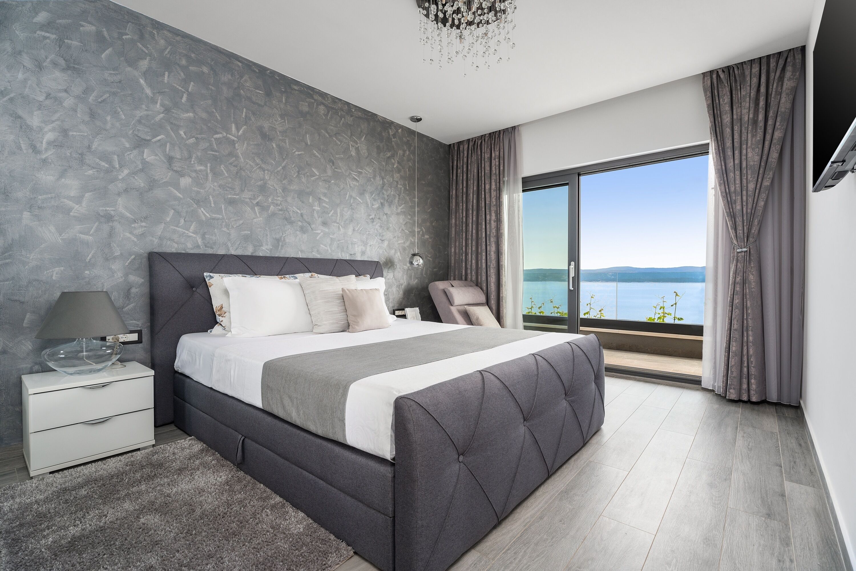 A modern bedroom with a large bed, a nightstand, a chair, and a view of the outdoors.
