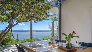 Une table à manger dressée avec de l’argenterie blanche, des verres et un saladier rempli de fruits, offrant une vue panoramique sur la mer.
