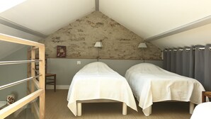 A small, cozy room with two single beds, a wooden chair, a small table, and a window with a view of the outside.