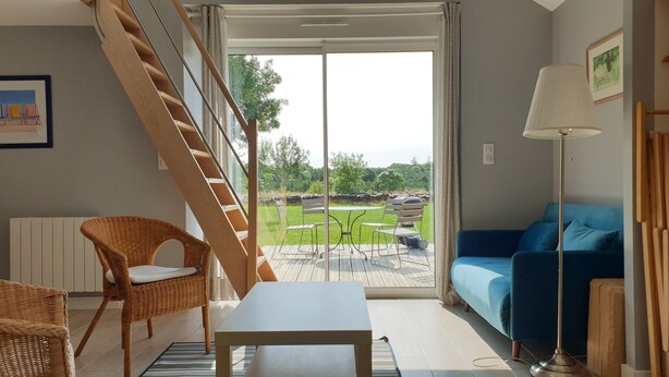 A living room with a staircase, a blue sofa, a wicker chair, a table, and a view of a garden through a glass door.