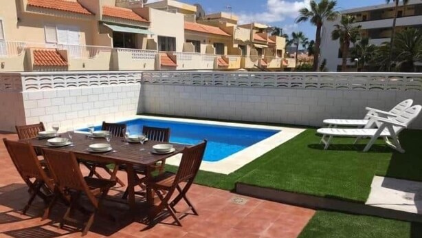 Una piscina con solárium empedrado, una mesa de comedor con sillas y una pequeña zona ajardinada con sillones de descanso.