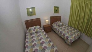 Una habitación con dos camas individuales, una mesita de noche con lámpara y una cortina verde.