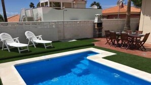 Una piscina con sillas de descanso blancas y un área de comedor con sillas de madera.