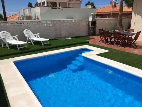 A swimming pool with white lounge chairs and a dining area with wooden chairs.