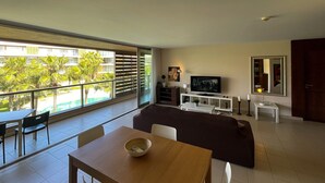 A modern living room with a large window, a flat-screen TV, a dining area, and a sofa. - Herdade dos Salgados, T1-10A-2D - Pool View, (Albufeira)