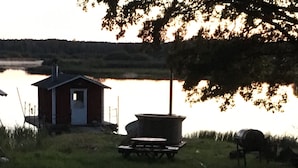 Una cabaña pequeña con puerta y ventanas, una estructura de piedra grande y una mesa de picnic cerca del agua.