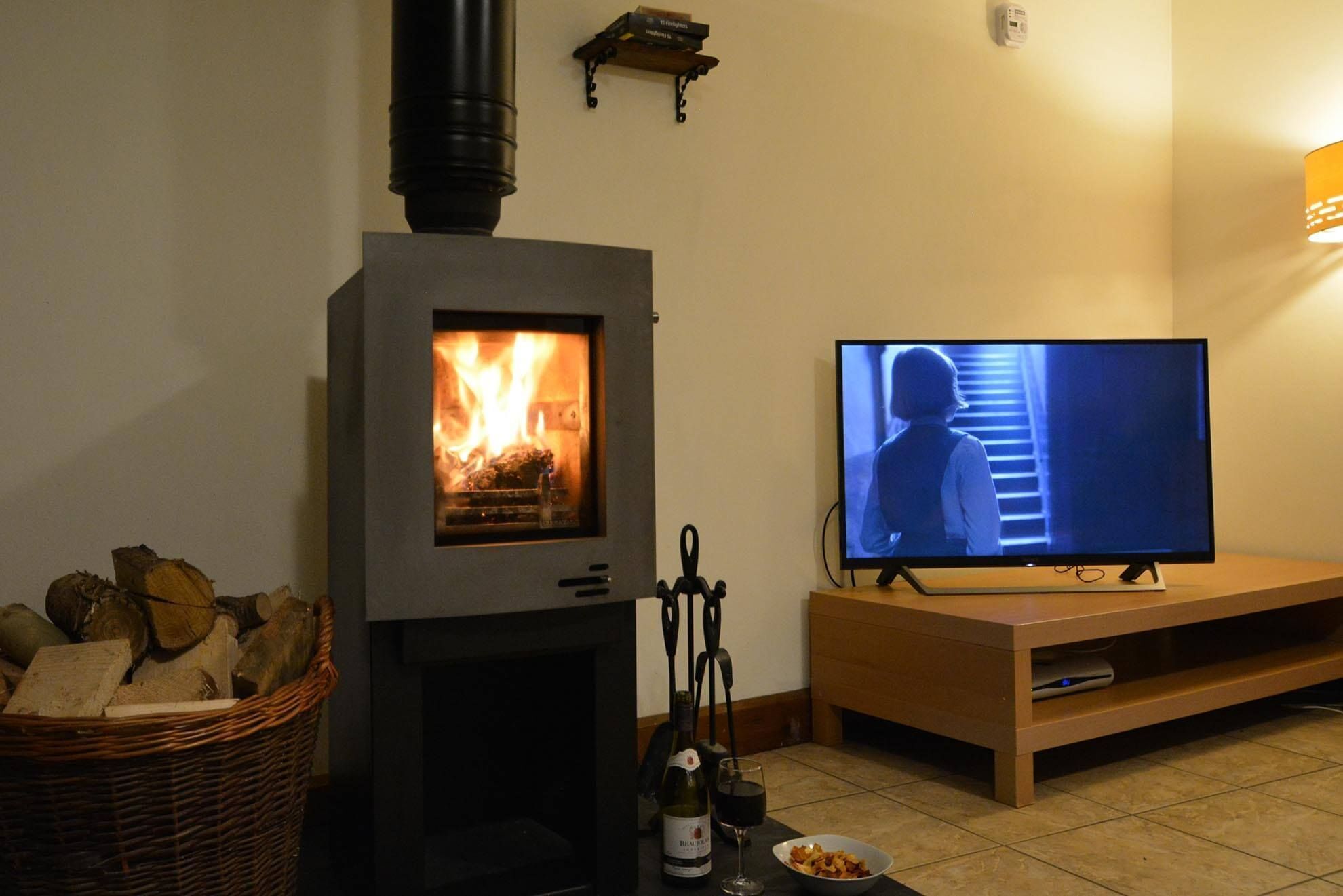 A living room with a wood-burning stove, a television, a wine bottle, and a basket of firewood.
