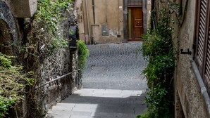 Une ruelle étroite sur laquelle passe un pont de pierre, bordée de bâtiments aux volets en bois et ornés de plantes en pot.