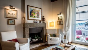 Un salon chaleureux avec une cheminée, deux fauteuils blancs, une table basse avec des livres et une vue sur un balcon fleuri.