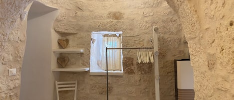 A rustic room with a stone wall, a window with a white curtain, a white chair, and a drying rack.