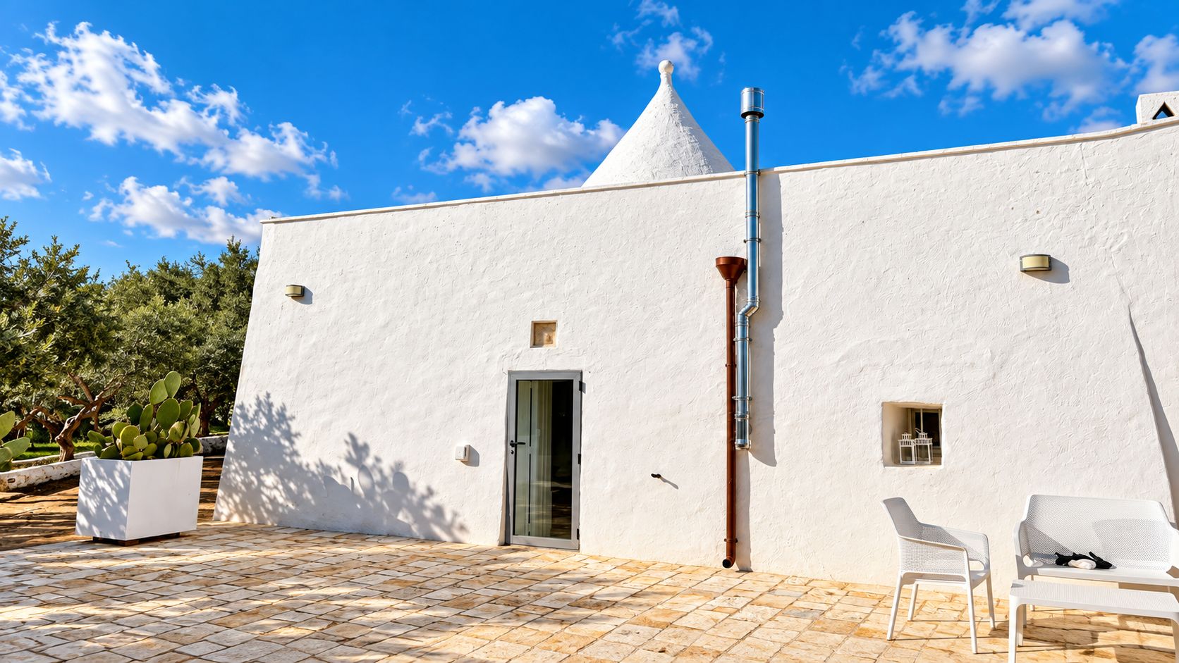 Un edificio blanco con una puerta, una ventana pequeña y una chimenea. Hay un patio con baldosas, sillas y una planta en maceta.