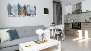 Un soggiorno moderno con un divano grigio, un tavolino da caffè bianco e un'area pranzo con tavolo e sedie bianche. Sullo sfondo, si intravede una cucina con forno a microonde e piano cottura.