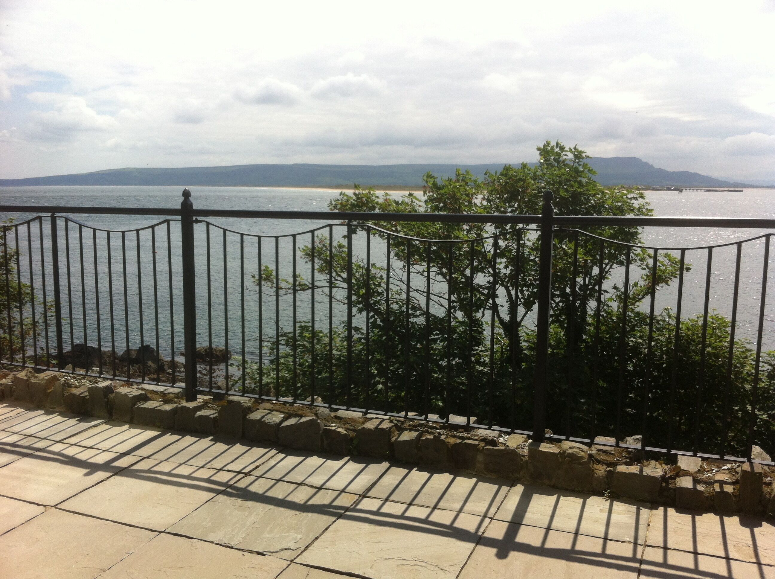 A stone-paved walkway with a metal railing overlooking a body of water and distant mountains.