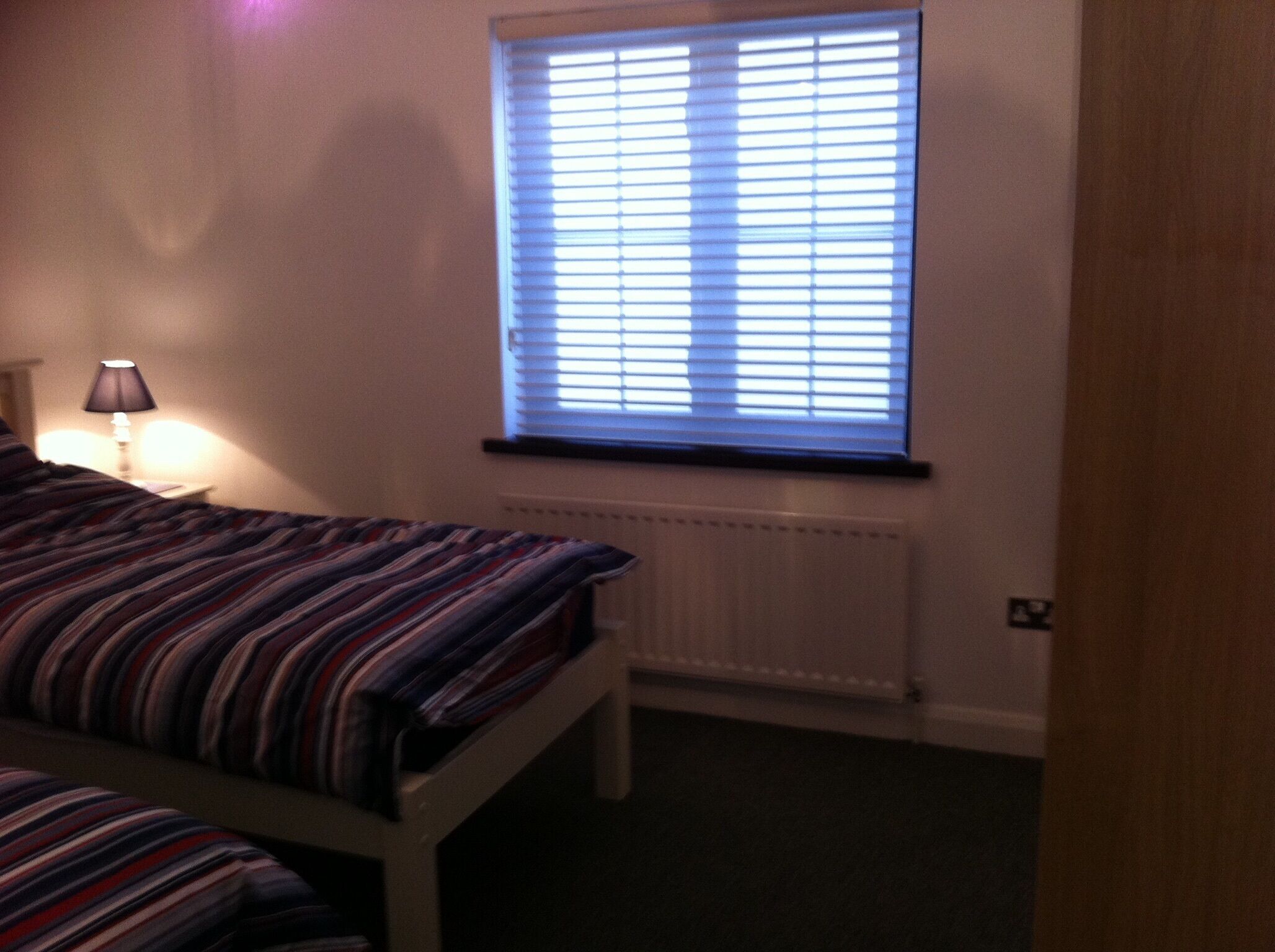 A bedroom with a bed, a window with blinds, a lamp, and a radiator.
