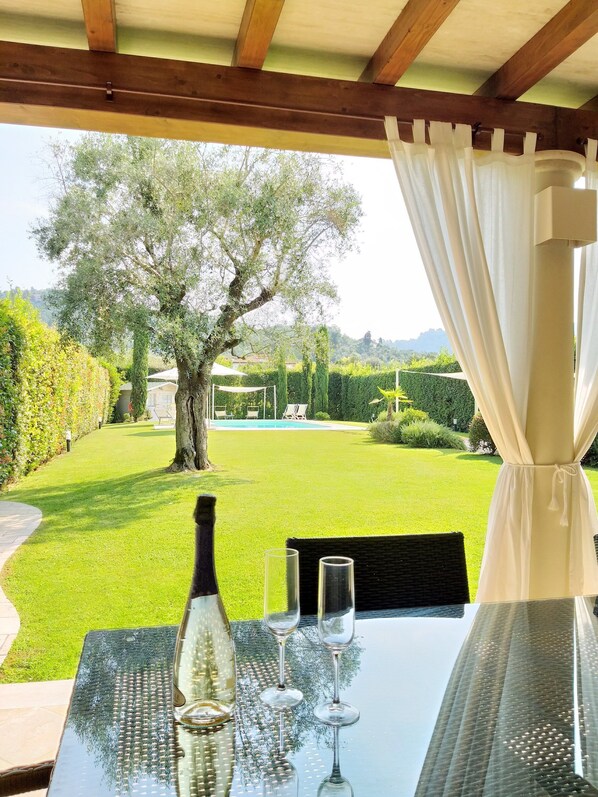 Una mesa con una botella y vasos, vista a un árbol y un área verde con una piscina al fondo.