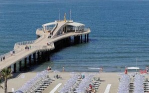 Un muelle que se adentra en el mar, con una plataforma de observación y una zona de playa con filas de tumbonas.