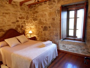 A bedroom with a stone wall, a wooden bed frame, a bedside lamp, and a window with wooden shutters.