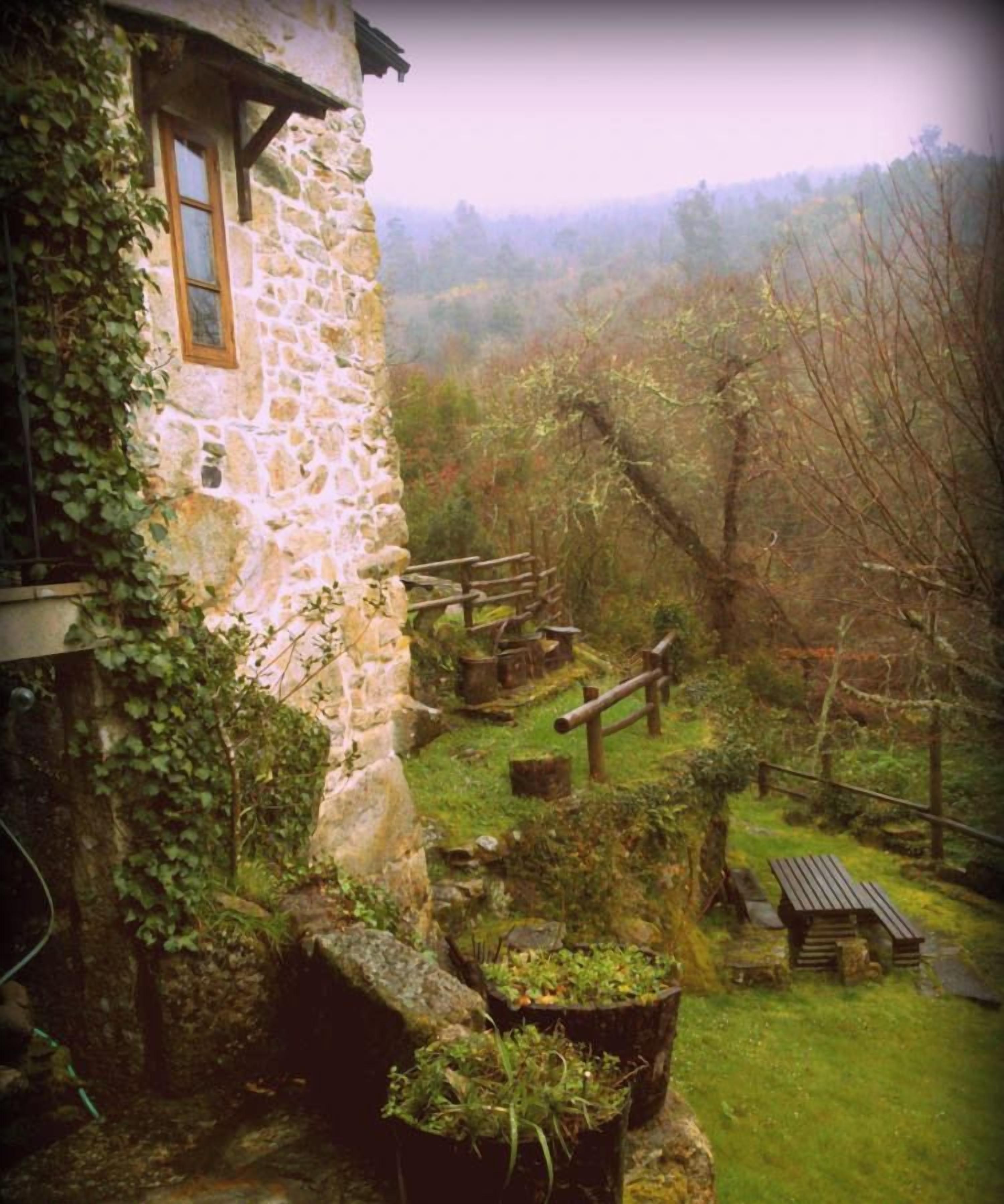 Un edificio de piedra con una ventana e hiedra, rodeado de césped, un banco de madera y una mesa de picnic.