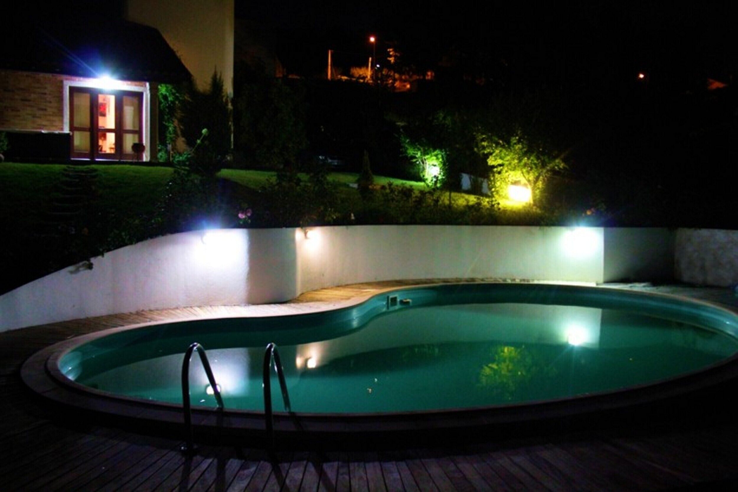 Une piscine éclairée la nuit, avec une terrasse en bois, entourée d’un petit muret et baignée par la lumière de lampes situées à proximité.