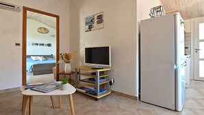 A compact living space with a bed, a television on a stand, a refrigerator, and a small table with magazines.