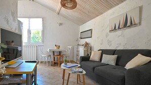 A cozy living room with a grey sofa, a wooden dining table, and a white chair. There's a window with a view of greenery outside.