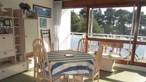 Een eethoek met een tafel en stoelen, een raam met uitzicht op bomen, en een boekenplank met diverse spullen.