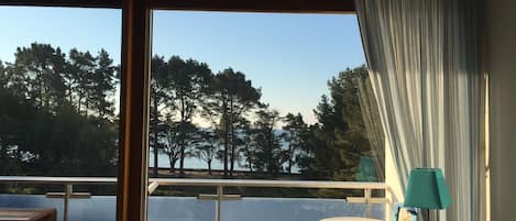 A room with a large window overlooking a body of water and trees. There is a chair and a small table with a lamp and a picture frame on it.