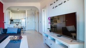 Ein modernes Wohnzimmer mit einer Couch, einem Couchtisch und einem Fernseher. Im Hintergrund ist eine Küche zu sehen.