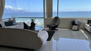 Una sala de estar moderna con un ventanal grande que da al mar, un sofá blanco, un sillón con estampado de cebra y una pequeña mesa con una planta.