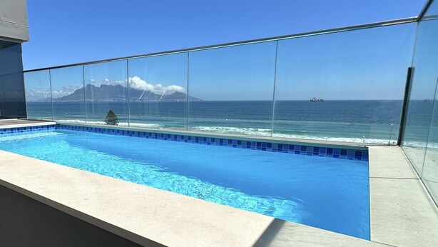 Una piscina con barandilla de cristal que domina el océano y una montaña al fondo.