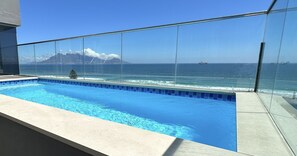 Piscina com guarda-corpo de vidro com vista para o oceano e uma montanha ao longe.