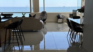 Una sala de estar moderna con un ventanal grande con vistas al mar, un sofá, una mesa de comedor con sillas y una pequeña zona de cocina.