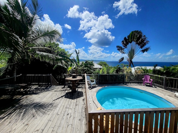 A wooden deck with a pool, surrounded by tropical plants and a view of the ocean.