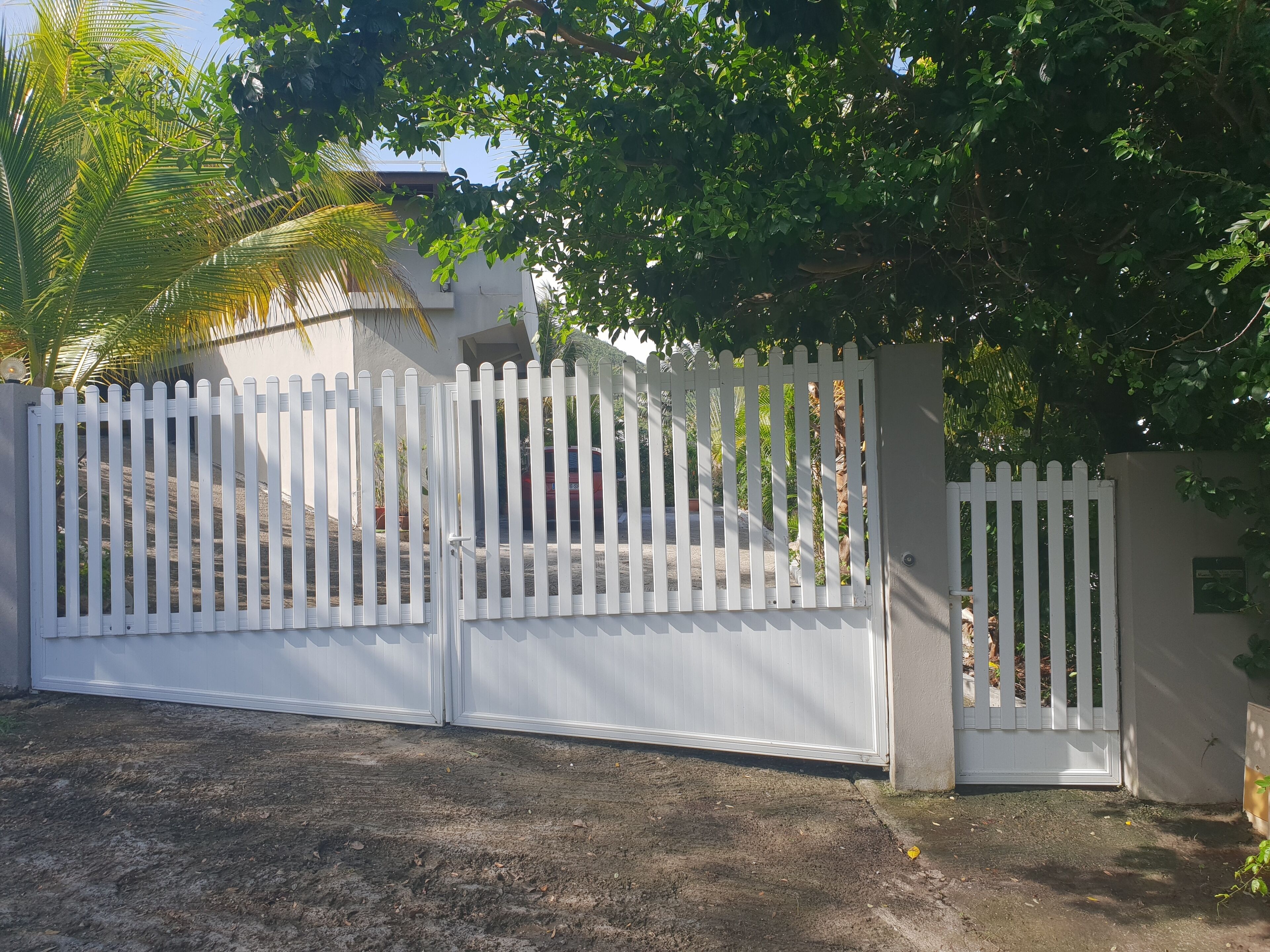 Une entrée fermée par un portail, avec une clôture en bois peinte en blanc, un mur en béton et un chemin pavé menant à un bâtiment.