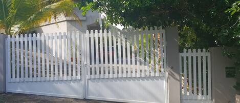 Une entrée fermée par un portail, avec une clôture en bois peinte en blanc, un mur en béton et un chemin pavé menant à un bâtiment.