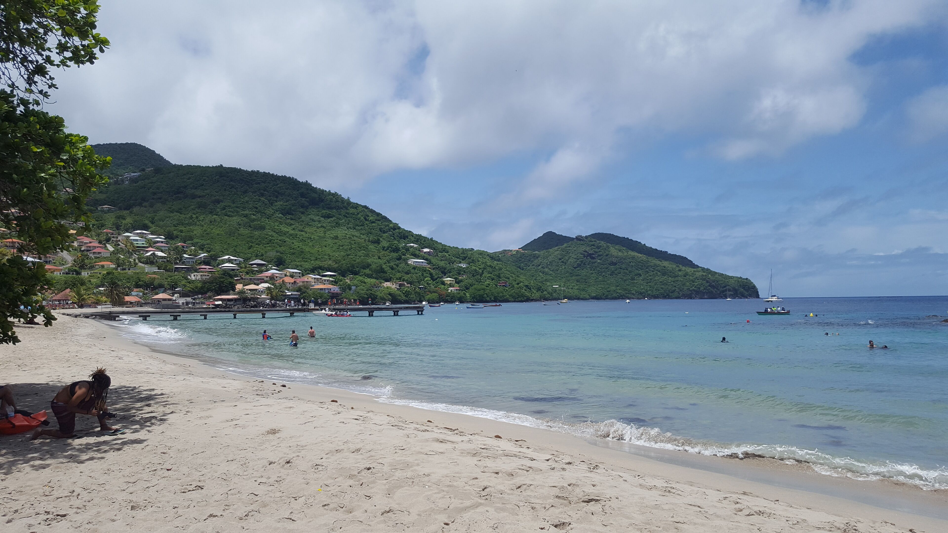 Ein Strand mit klarem Wasser, einer Mole und Gebäuden an einem Hügelhang.