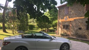 A white convertible car parked on a gravel driveway in front of a stone building with a circular emblem on the wall. - ancient villa with pool and park in total privacy (tuoro sul trasimeno)